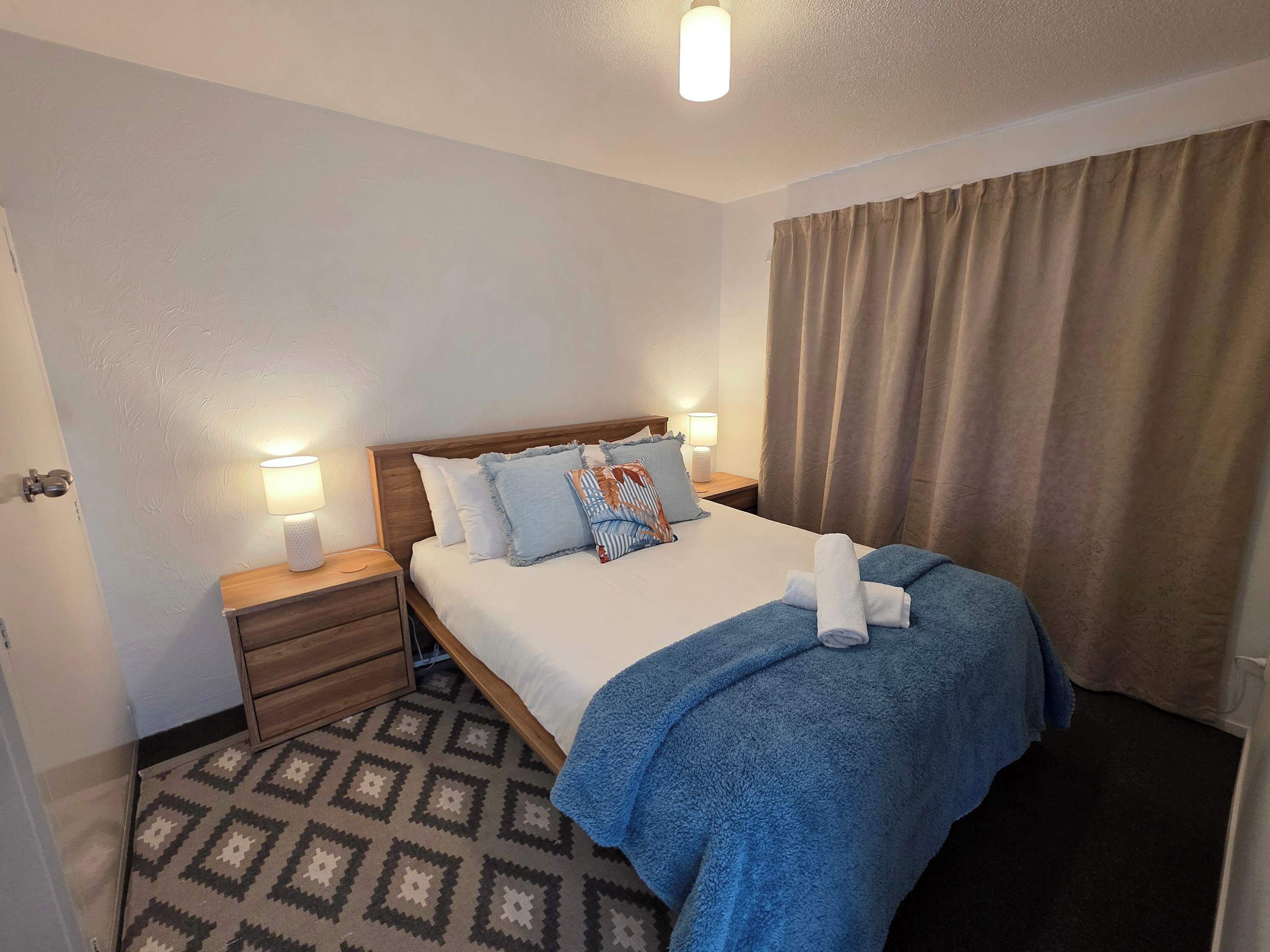 Een hotelkamer met een bed, nachtlampjes, een nachtkastje, een kamerbreed tapijt met een patroon en een raam met gordijnen.