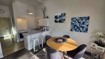 A modern kitchen with white cabinets, a dining area with a round wooden table and grey chairs, and a living space with a sofa and decorative items.