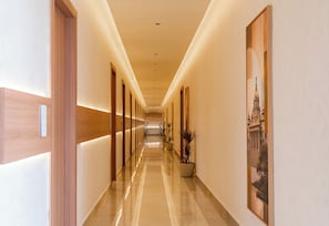A hotel corridor with wooden doors, a polished floor, and framed artwork on the walls. - ALAYA STAYS SAVIAN Suites (Chikkajala)