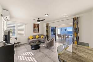 Een moderne woonkamer met een bank, fauteuil, salontafel en een televisie. Er is een balkon met uitzicht op de zee.