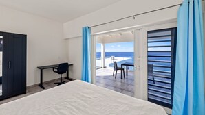 Un dormitorio con cama, escritorio y silla, un armario y un balcón con vista al mar.