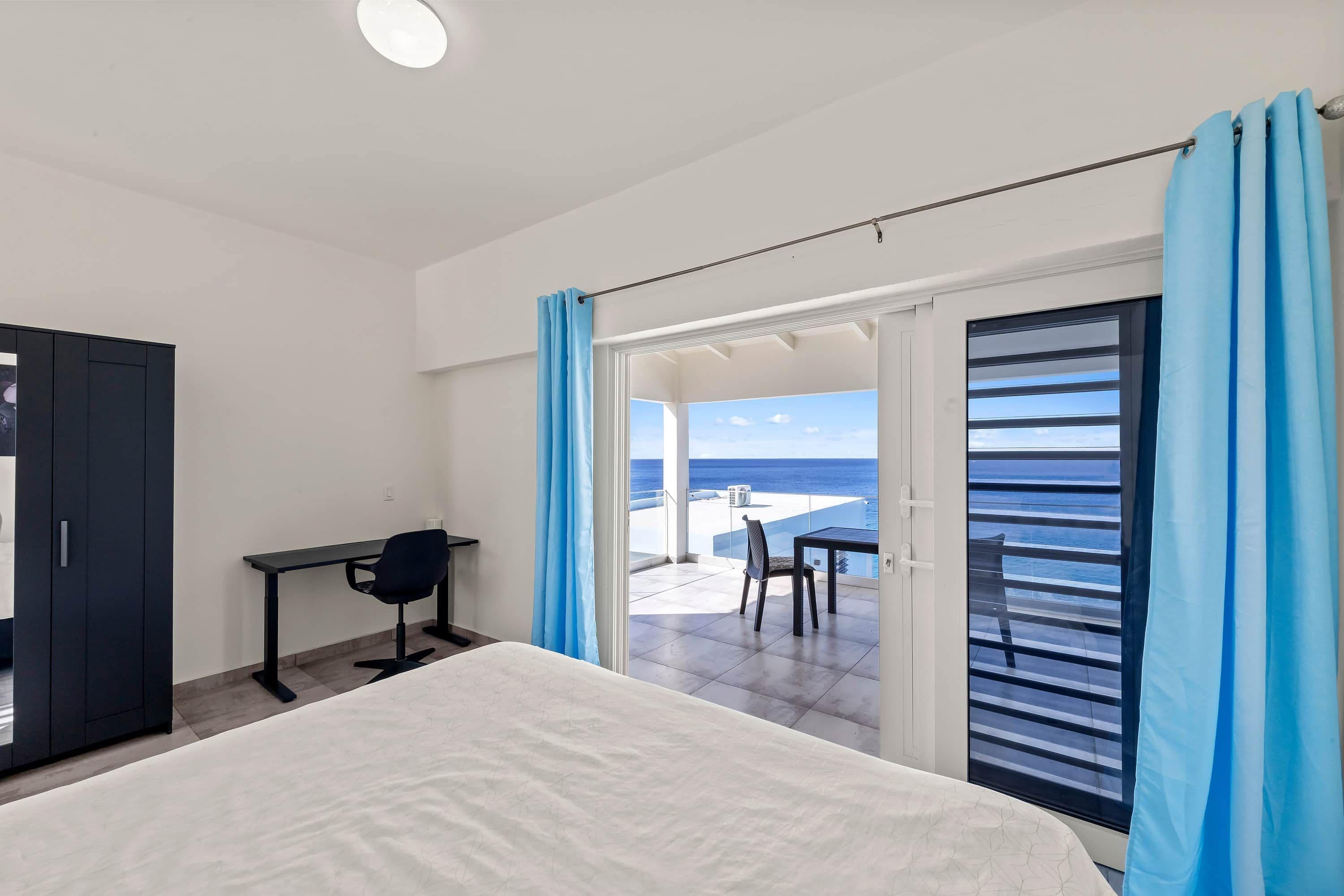 Une chambre avec un lit, un bureau et une chaise, une armoire et un balcon donnant sur la mer.