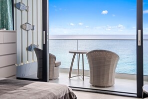 Ein Balkon mit Meerblick, zwei Korbstühle, ein kleiner Tisch und ein Bett mit grauem Kopfteil.