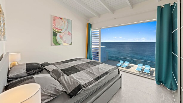 Un dormitorio con cama, un cuadro, una mesita de noche, una lámpara y vista al mar desde un balcón.