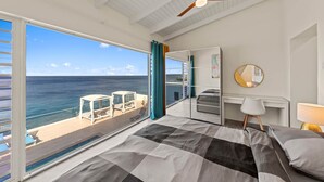 Un dormitorio con una ventana grande, un ventilador de techo, una cama, un escritorio y vistas al océano.