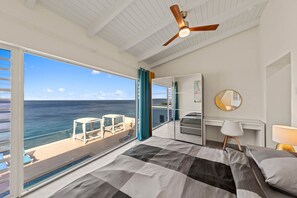 Ein Schlafzimmer mit großem Fenster, Deckenventilator, Bett, Schreibtisch und Blick auf den Ozean.