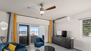Una sala de estar moderna con un sofá, una mesa redonda de centro, ventilador de techo, televisor y balcón con asientos exteriores.