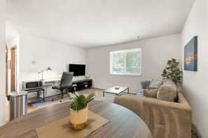 A modern living room with a dining area, a flat-screen TV, a desk with a computer, and a large window with blinds. - Near UW Campus | Parking & Pet-Friendly (Madison)