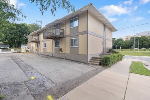 Ein mehrstöckiges Gebäude mit einem Balkon im zweiten Stock, einem Parkplatz davor und einem Bürgersteig mit einer Grünfläche zur Rechten.