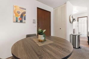 A round dining table with a plant centerpiece, a wall-mounted abstract painting, and a closed door with a louvered window. - Near UW Campus | Parking & Pet-Friendly (Madison)