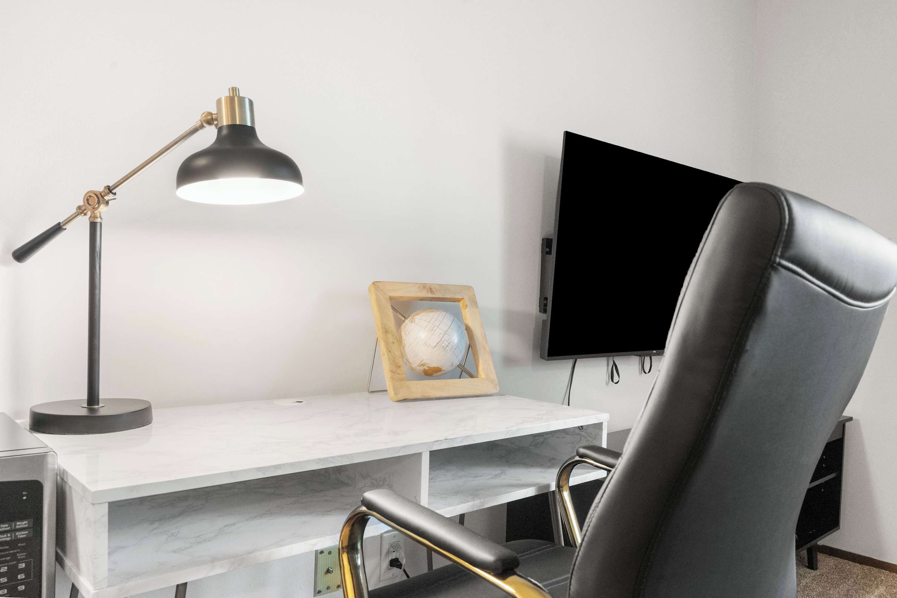 A modern office chair, a desk with a lamp, a framed picture, and a flat-screen TV mounted on the wall.