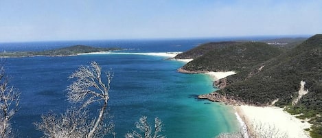 A coastal landscape with a beach, clear blue waters, and forested hills.