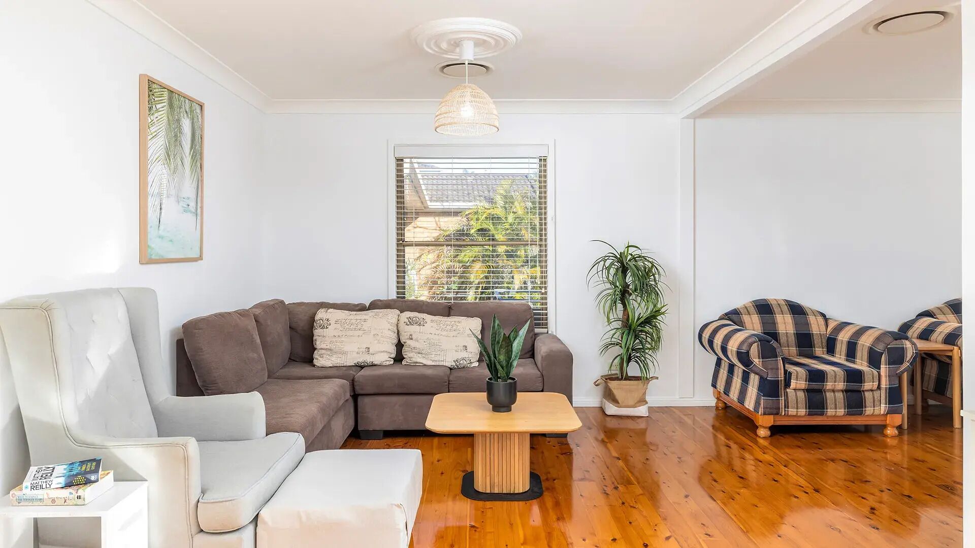 A living room with a sectional sofa, a white armchair, a plaid sofa, a wooden coffee table, and a potted plant.