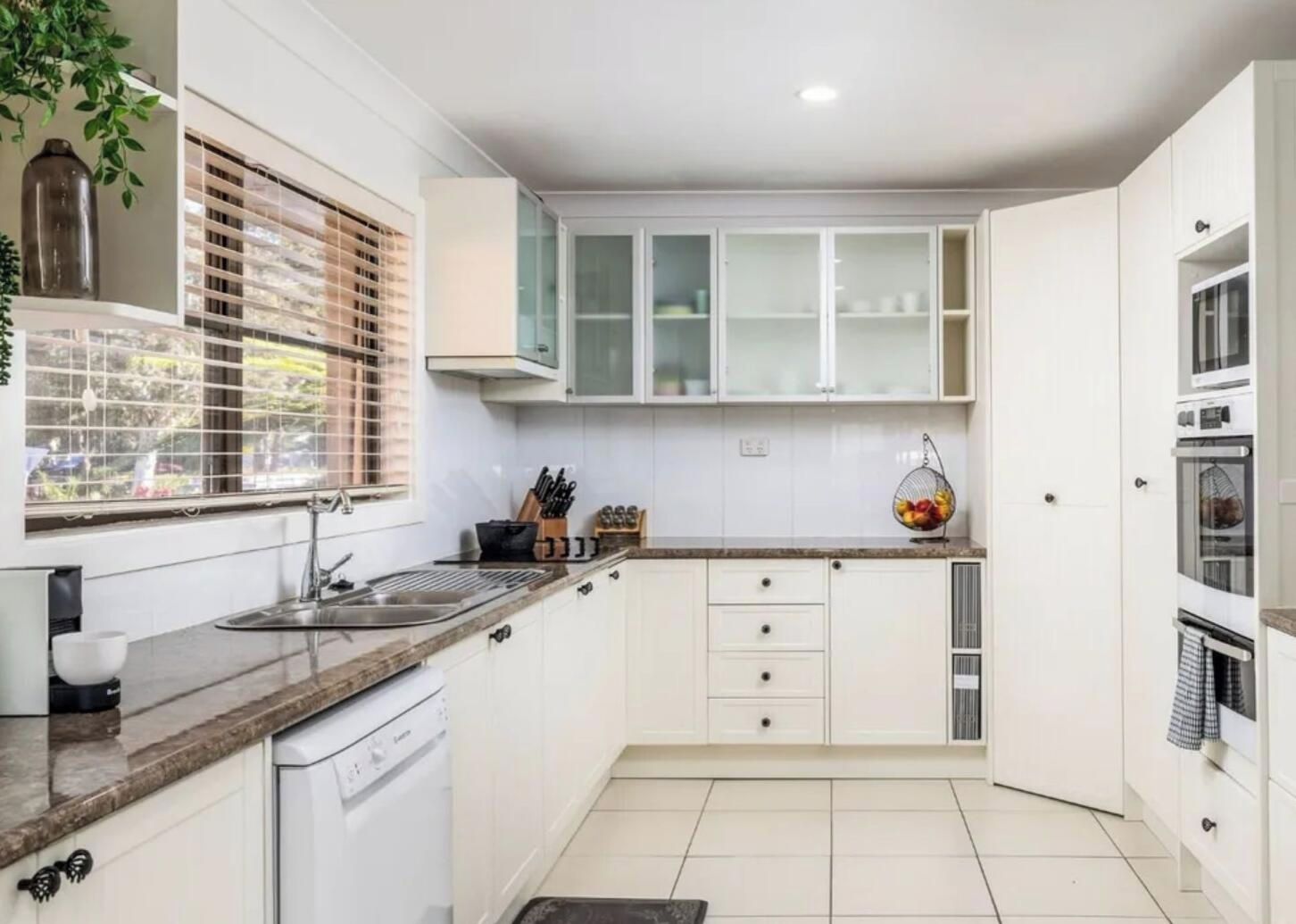 A modern kitchen with white cabinets, a dishwasher, and a window with blinds.