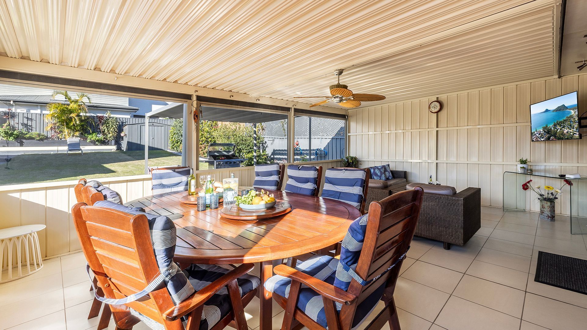 A covered outdoor dining area with a wooden table and chairs, a ceiling fan, and a view of the backyard.