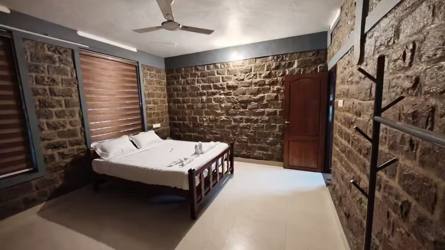 Quarto com cama, ventilador de teto, paredes de pedra e porta de madeira.