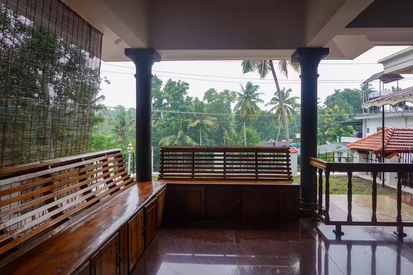 A covered balcony with wooden benches and a railing, overlooking a residential area with palm trees.
