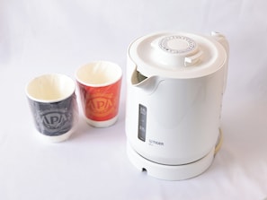A white electric kettle with a digital display and a lid, next to two mugs with logos. - APA Hotel Shibuya Ekimae (Tokyo)