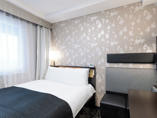 A hotel room with a bed, a desk, a chair, and a nightstand with a lamp.