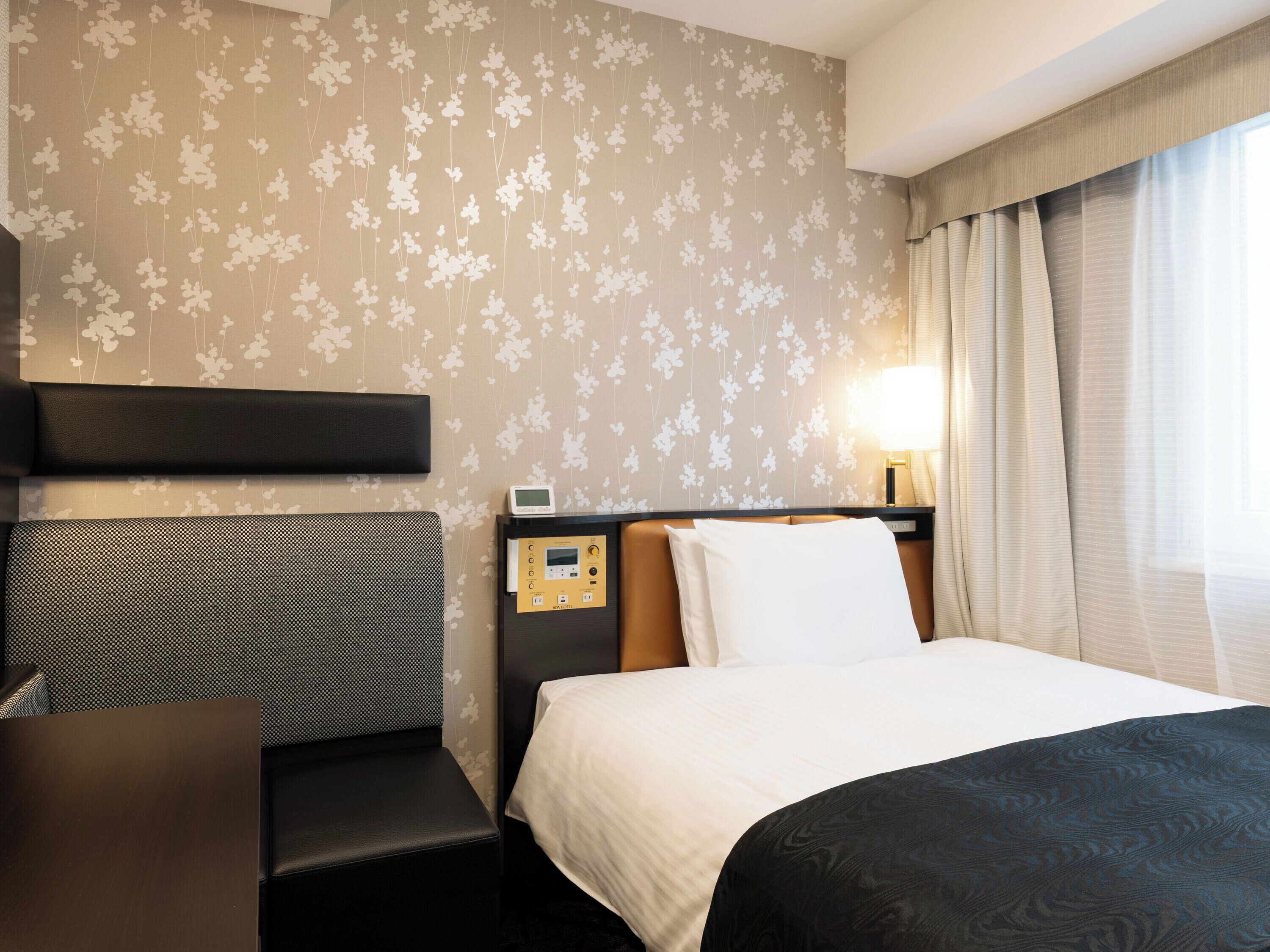 A hotel room with a bed, a desk, a nightstand, and a wall-mounted control panel.