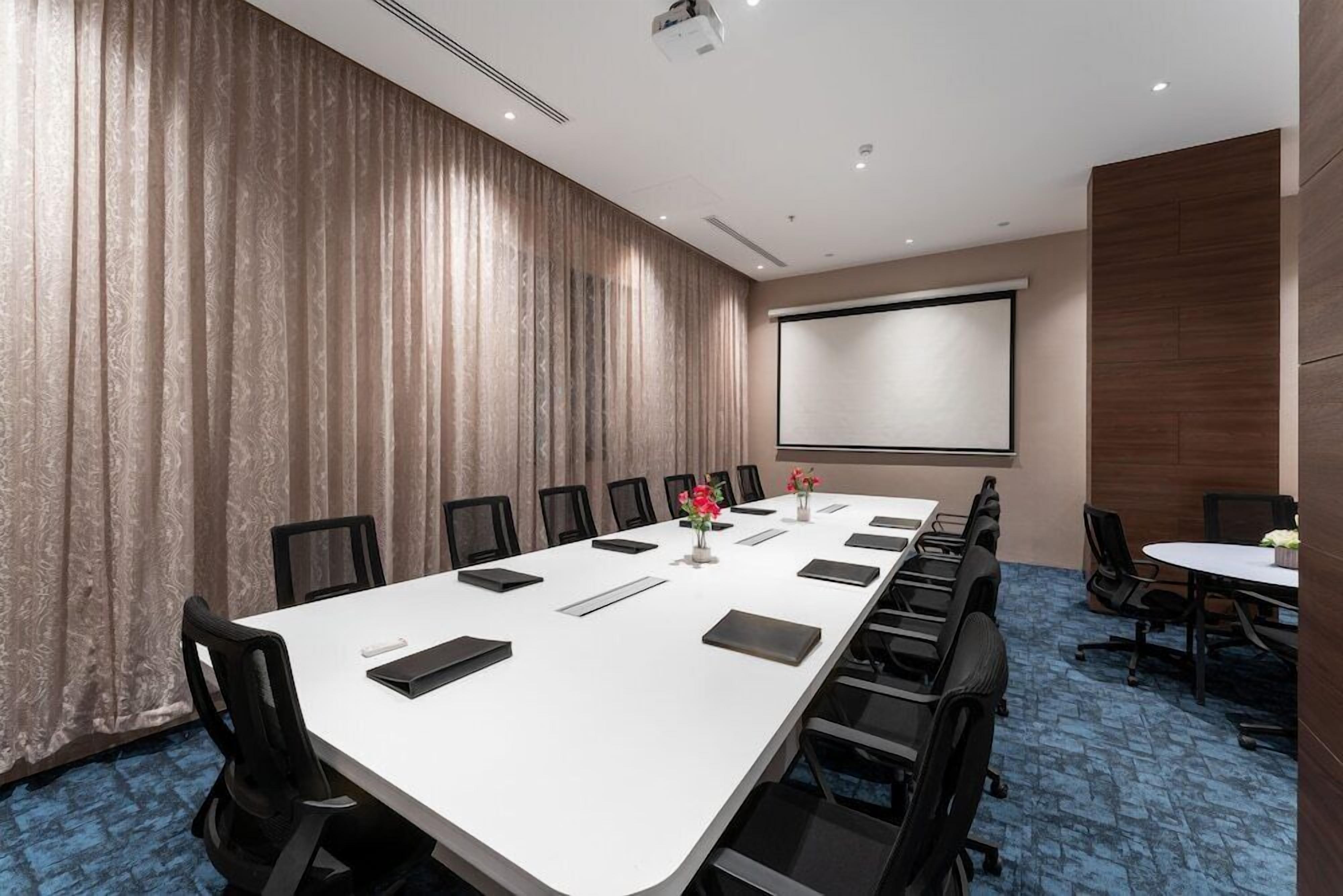 A conference room with a long table, chairs, a projector screen, and a blue carpeted floor.