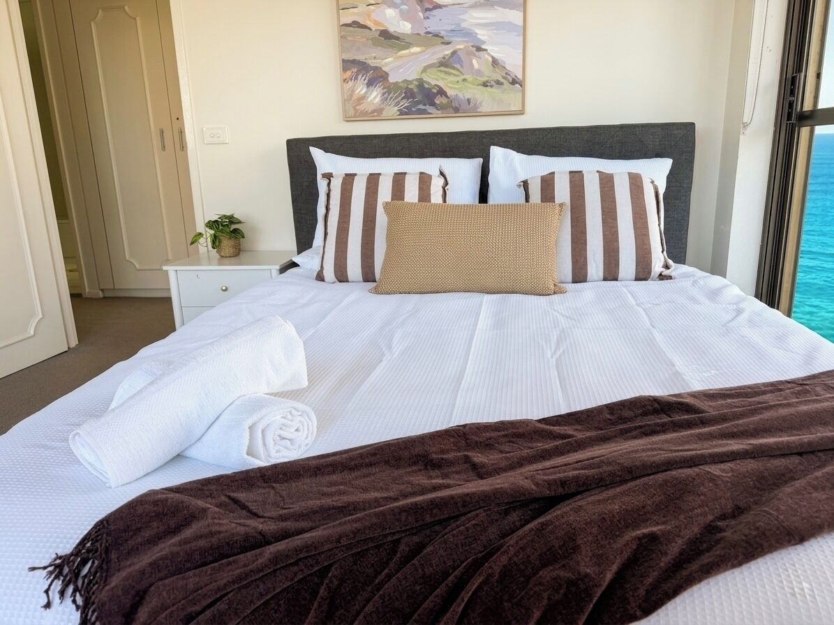 A neatly made bed with white linens and a brown throw blanket, a bedside plant, and a painting on the wall.