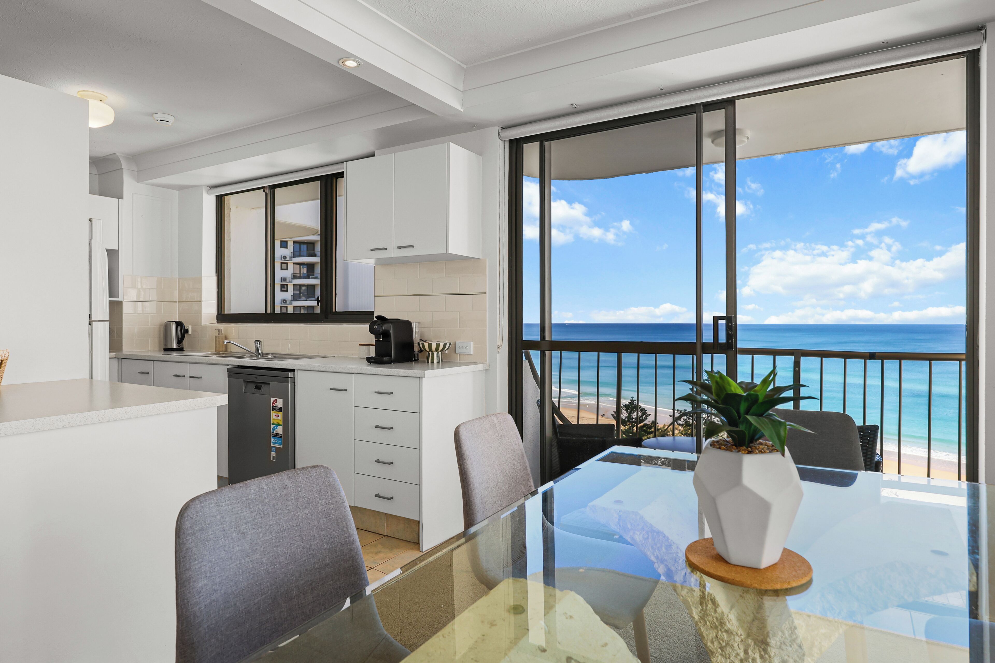 A modern kitchen with a view of the ocean, a dining area with a glass table, and a balcony with a view of the beach.
