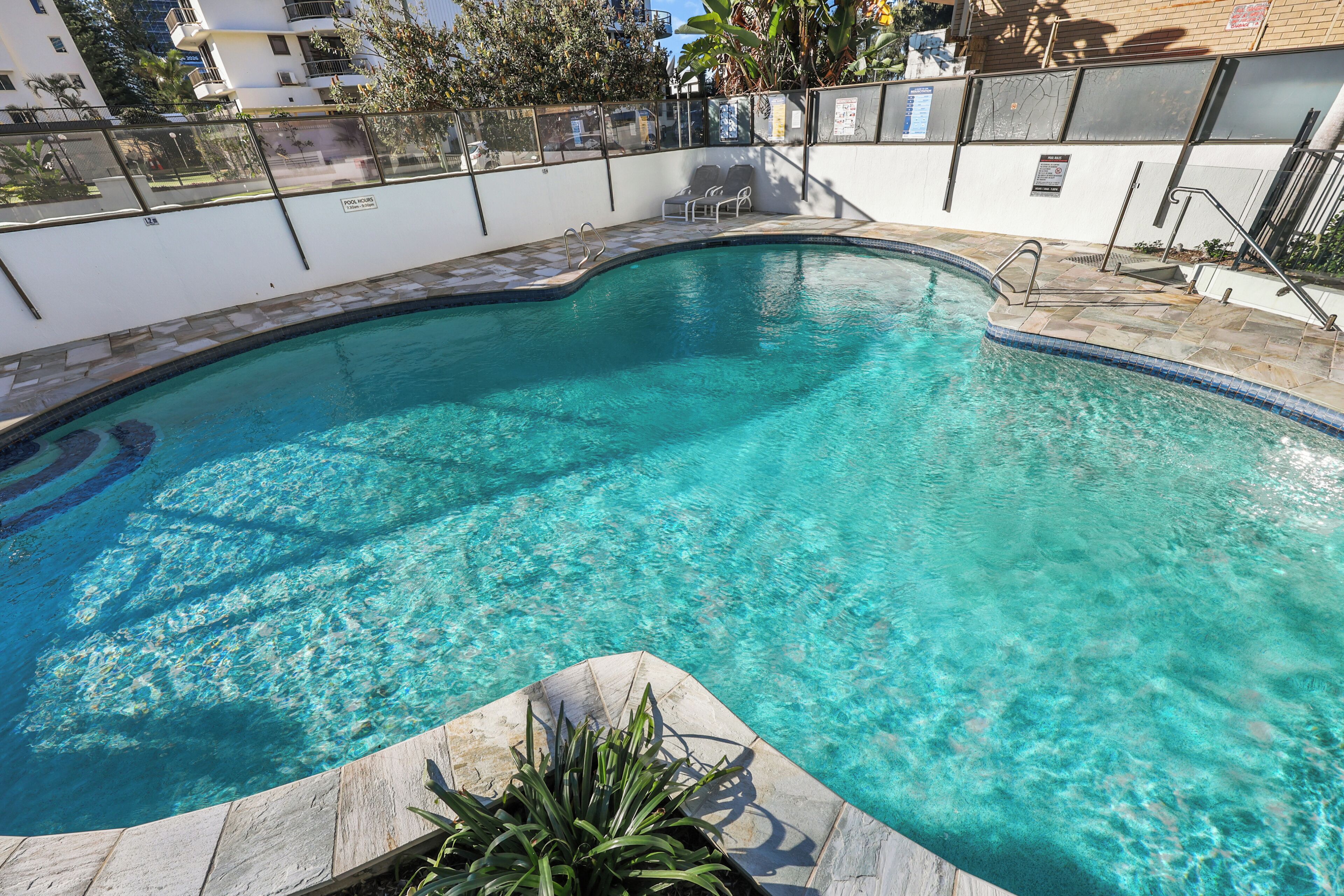 A clear swimming pool with a curved edge, surrounded by a low wall and a metal fence.