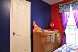 A bedroom with a wooden dresser, a bed with a colorful quilt, a door, and a window with purple curtains.