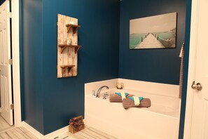 A bathroom with a bathtub, a wooden shelf, a picture of a pier, and a small basket. - The "Home Over the Bridge" (Atmore)