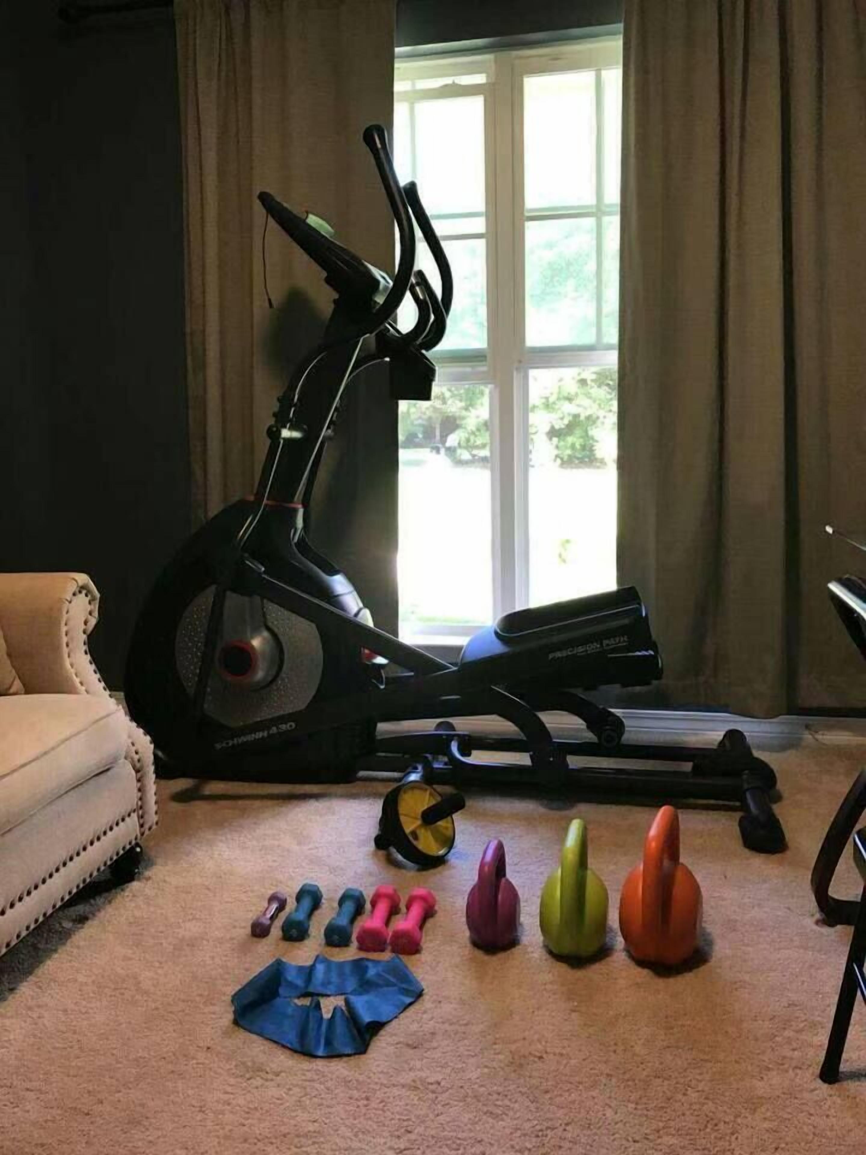Une salle de sport à domicile équipée d’un vélo elliptique, d’un tapis de course et de divers haltères de couleurs différentes.