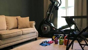 A living room with a beige sofa, a stationary exercise bike, and kettlebells on the floor.