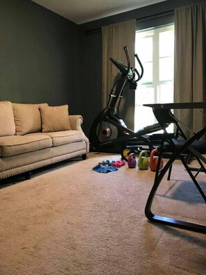 A living room with a beige sofa, a stationary exercise bike, and kettlebells on the floor.