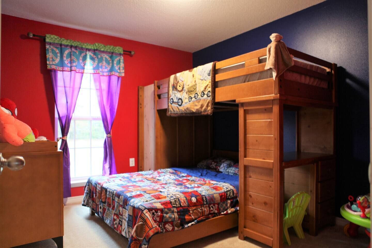 A bunk bed with a colorful quilt, a window with purple curtains, and a toy chair.