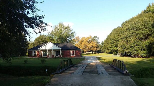 A house with a front porch, a driveway leading to a wooden bridge, and a well-maintained lawn.