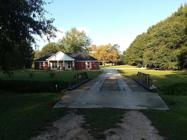 A house with a front porch, a driveway leading to a wooden bridge, and a well-maintained lawn.