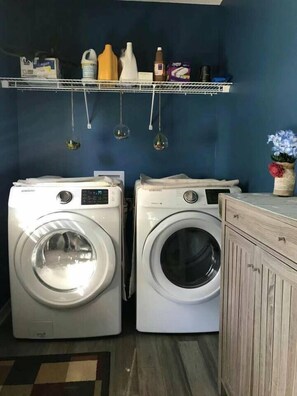 A laundry room with two front-loading washing machines, a shelf with cleaning supplies, and a wooden cabinet.