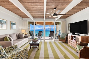 A living room with a sofa, armchairs, a coffee table, and a television. There is a balcony with a view of the ocean.