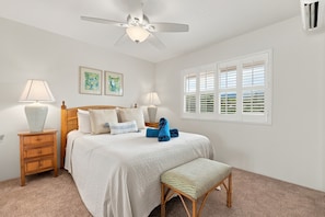 A bedroom with a bed, two bedside lamps, a ceiling fan, a window with shutters, and two framed artworks on the wall.