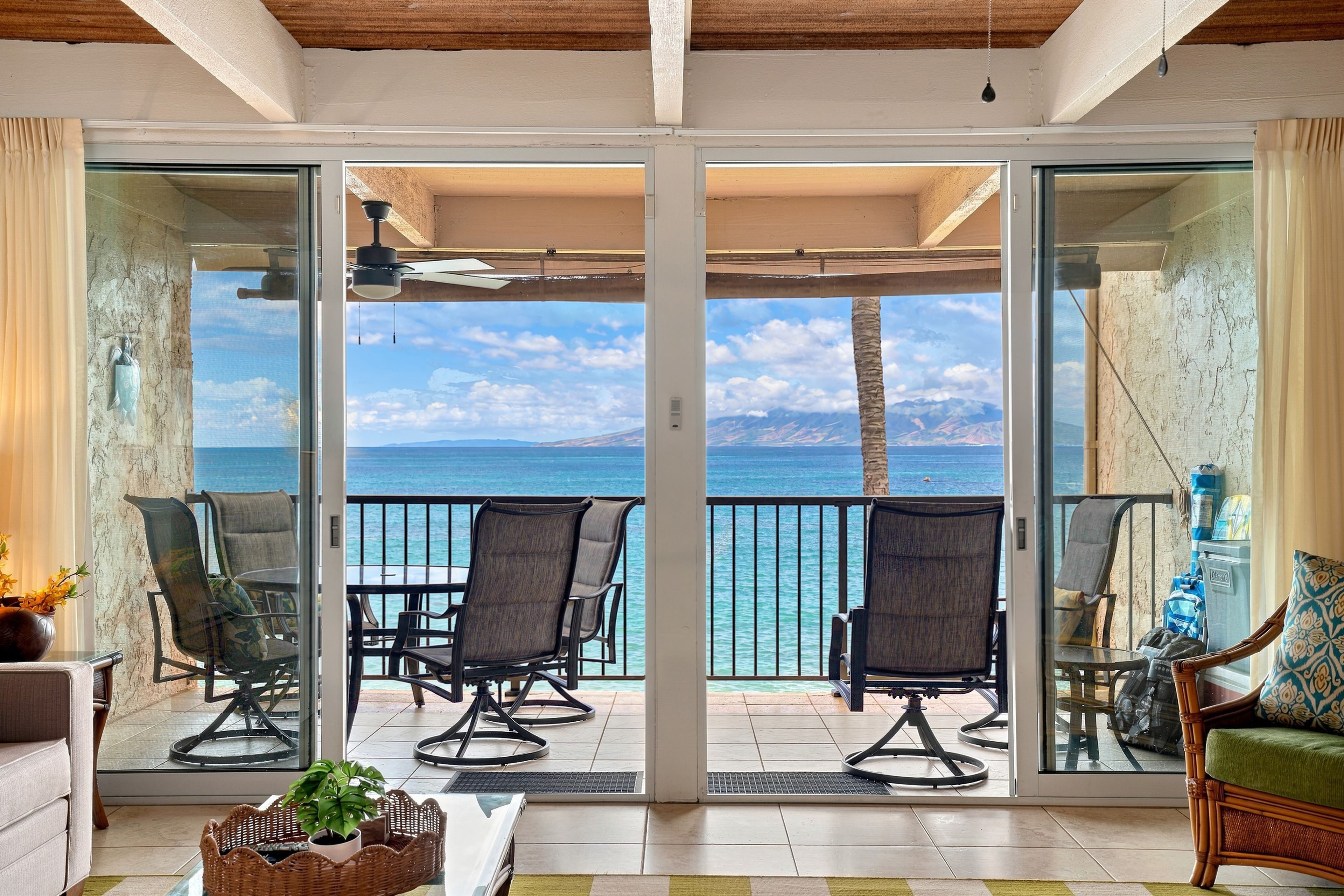 A room with a sliding glass door leading to a balcony with a view of the ocean and mountains. There are chairs and a table on the balcony.
