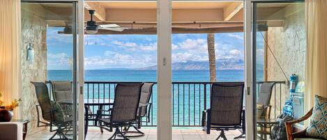 Quarto com porta de vidro deslizante que dá para uma varanda com vista para o oceano e montanhas. Há cadeiras e uma mesa na varanda.