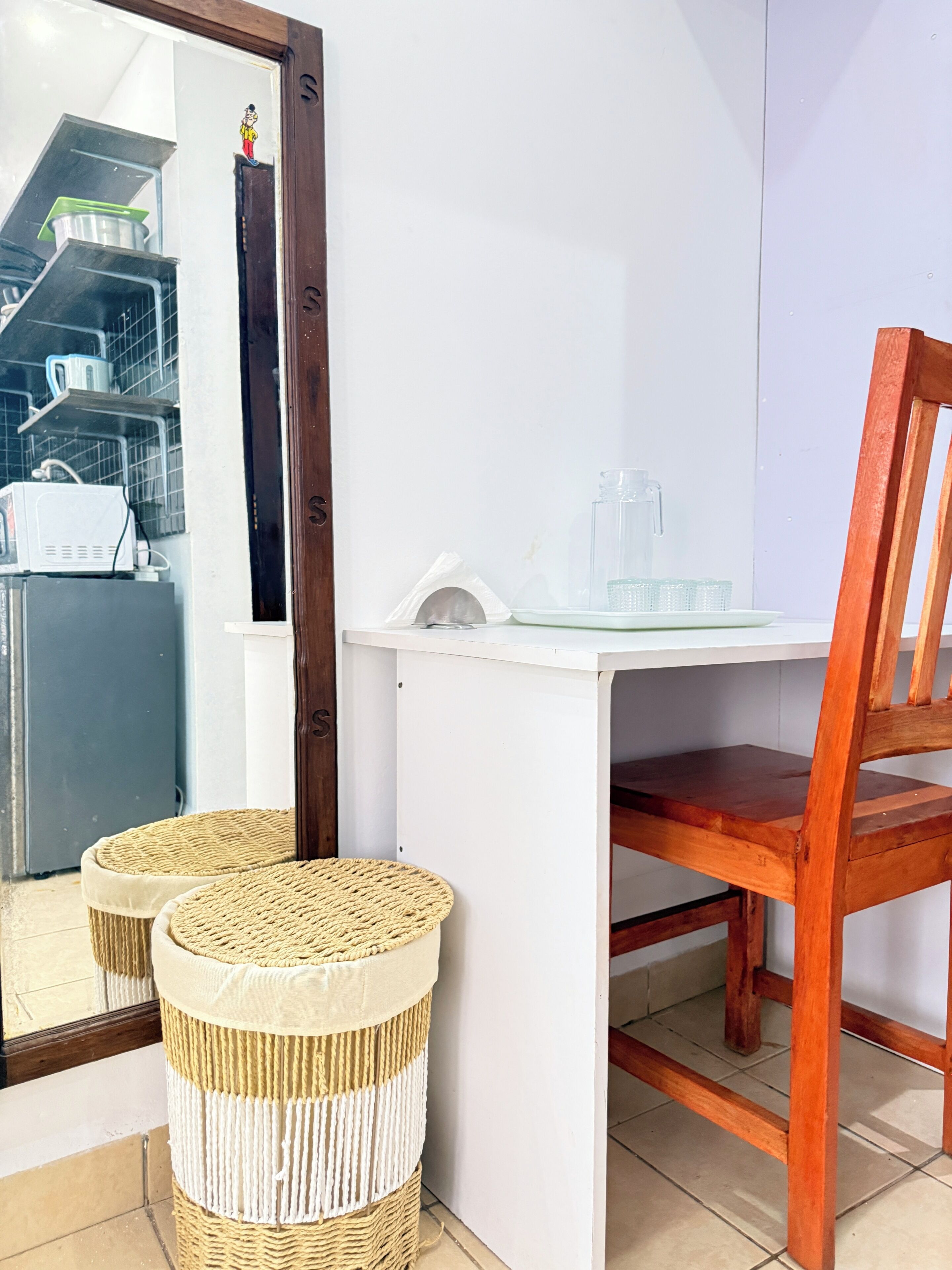 A room with a white desk, a wooden chair, and two woven stools. A mirror reflects a kitchen area with shelves and appliances.