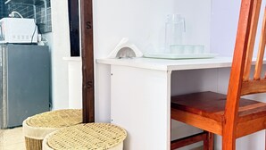 Quarto com escrivaninha branca, cadeira de madeira e dois banquinhos trançados. Um espelho reflete uma cozinha com prateleiras e eletrodomésticos.