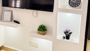 Uma TV na parede, um relógio, uma moldura com uma frase, um controle remoto e duas plantas em vasos.