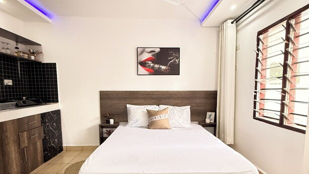 Quarto com cama, ventoinha no teto, janela com persianas, área de cozinha com painel de azulejos pretos e um quadro pendurado na parede.