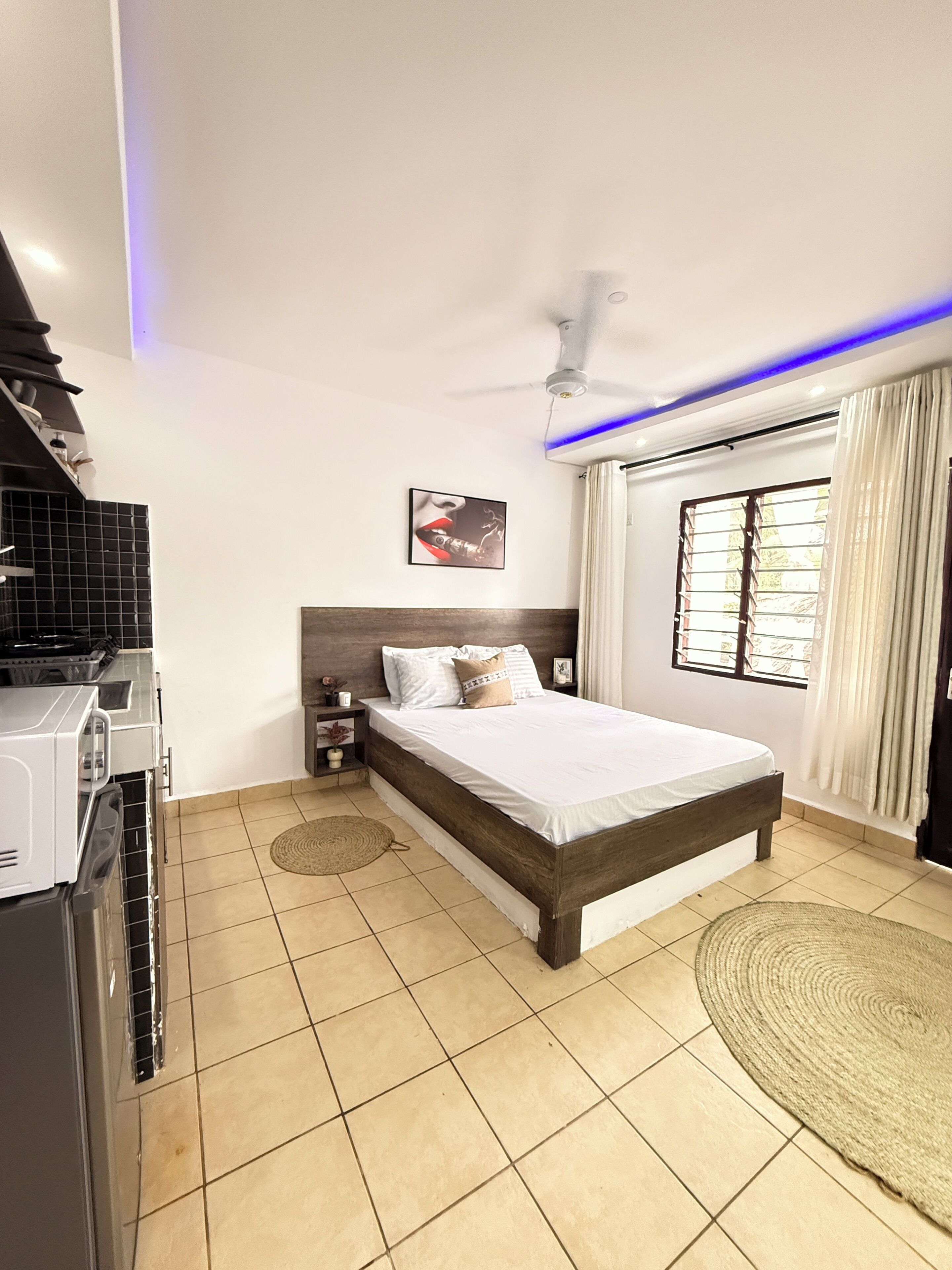 A bedroom with a bed, a ceiling fan, a window with curtains, and a kitchen area with a stove.