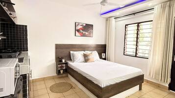 A bedroom with a bed, a ceiling fan, a window with curtains, and a kitchen area with a stove.