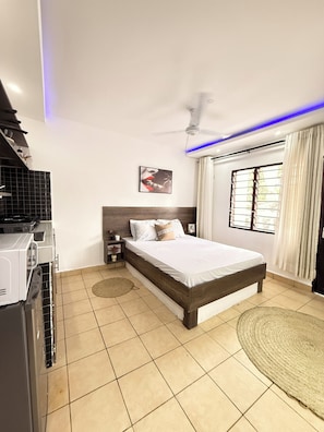 Ein Schlafzimmer mit einem Bett, einem Deckenventilator, einem Fenster mit Vorhängen und einer Küchenzeile mit einem Herd.