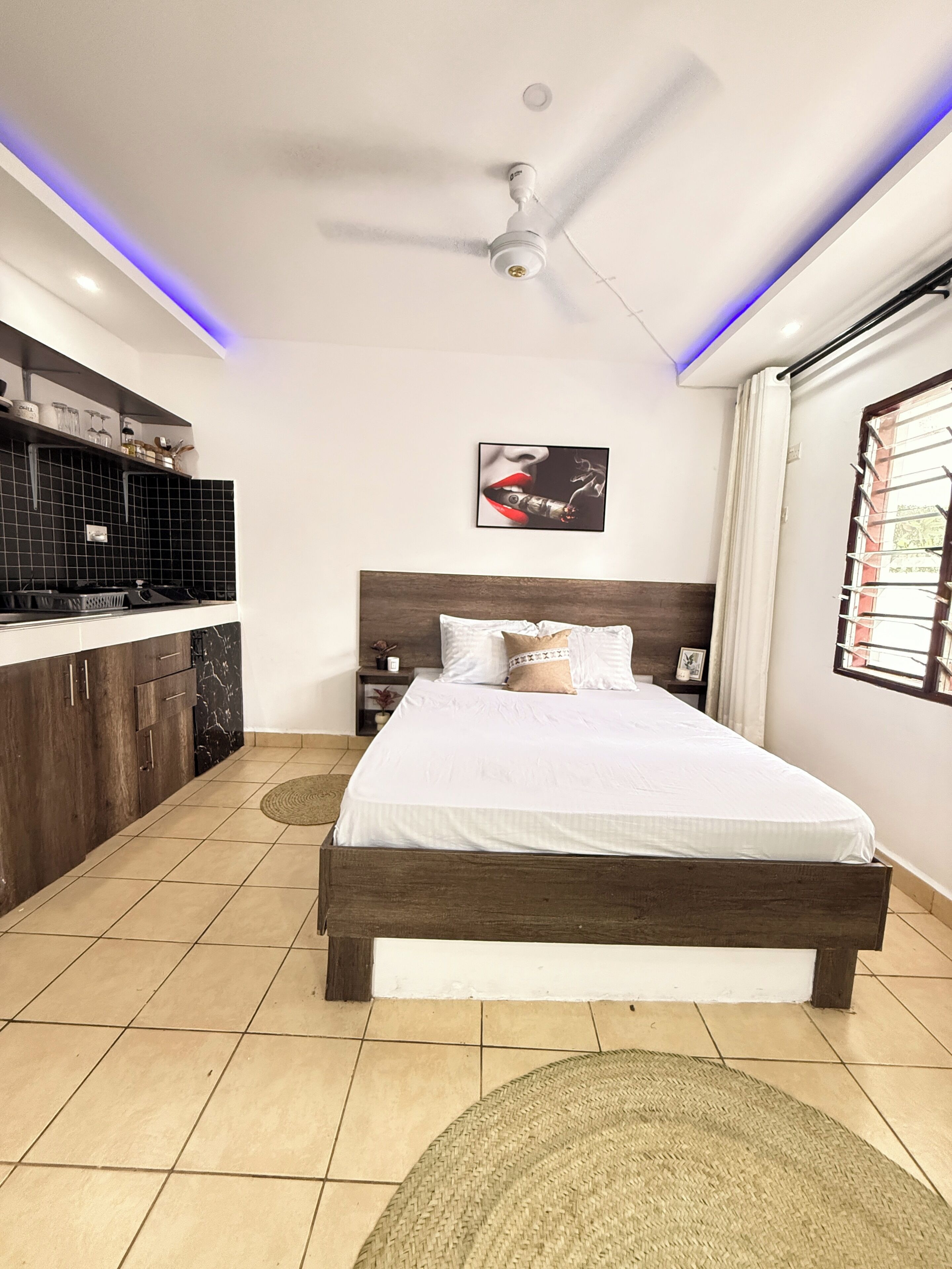 A bedroom with a bed, a ceiling fan, a window with curtains, a kitchen area with a black backsplash, and a wall-mounted picture.
