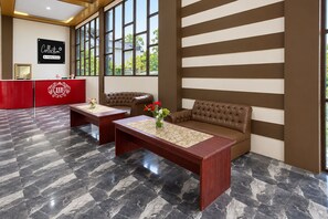 Un hall de hotel con una recepción, una zona de estar con un sofá y una mesa, y un amplio ventanal con vistas al exterior.